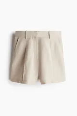 Short sastrero color beige de tiro alto, confeccionado en mezcla de lino. Presenta un diseño holgado con elástico en la parte posterior de la pretina, cierre oculto con botón y corchete, bolsillos al bies, pliegues delanteros y raya marcada en la parte delantera y trasera.