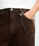 Pantalón de sarga color marrón oscuro, con corte super baggy, cintura con trabillas, bolsillos laterales y traseros, y cierre frontal con botón y cremallera. Incluye una cadena metálica decorativa en la cintura.