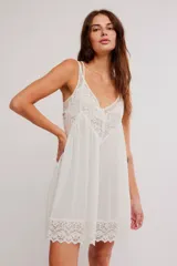 Vestido corto tipo slip dress color blanco, de corte amplio tipo babydoll, confeccionado en mezcla de viscosa y algodón. Presenta escote en V con apliques de encaje, tiras finas regulables y ruedo festoneado con detalles de encaje y calados.
