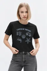 Remera negra de algodón con cuello redondo, manga corta y estampado frontal inspirado en temática marina con texto "Catch of the Day".