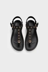 Sandalia baja de cuero vacuno color negro, tipo thong sandal, con tira vertical en el empeine decorada con apliques metálicos en forma de flores y tira ajustable en el talón con hebilla.