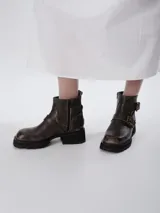 Bota corta de cuero color marrón, con suela track gruesa y taco cuadrado. Presenta un diseño con doble correa lateral con hebillas metálicas y cierre lateral para mayor comodidad.