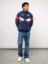 Buzo deportivo de nylon con medio cierre frontal y cuello alto. Presenta un diseño de bloques de color en azul, blanco y bordó, con el escudo del Club Nacional de Football y el logo de Umbro en el pecho.