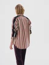Camisa de denim con estampado de rayas verticales en tonos bordó y crema. Presenta cuello clásico, cierre frontal con botones negros, canesú trasero negro y paneles laterales negros en las mangas. Incluye un pequeño bordado con el nombre de la marca en el pecho.