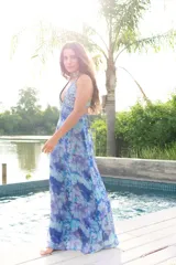 Vestido largo con estampado tie-dye en tonos violeta y fucsia, escote en V con detalles bordados dorados, cut-outs laterales bajo el busto y tirantes finos que se cruzan y atan en la espalda.