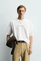 Camiseta blanca de corte relajado, con cuello redondo, manga corta y estampa frontal de texto pequeño en marrón.
