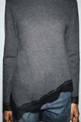 Top de manga larga en tejido suave color gris, con cuello redondo y bajo asimétrico decorado con encaje negro a tono. Presenta detalles de encaje negro en los puños.