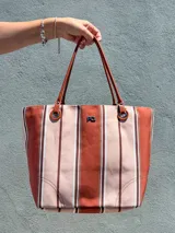 Cartera tipo tote bag confeccionada en canvas de algodón con diseño de rayas verticales en tonos beige, terracota y blanco. Cuenta con asas finas de cuero sintético marrón, detalles de argollas metálicas y logo metálico de la marca en el frente.