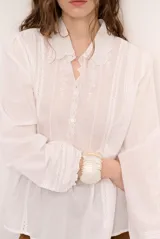 Camisa blanca de manga larga con detalles bordados en el frente, cuello redondo con solapas delicadas, cierre frontal con botones y puños anchos con encaje.