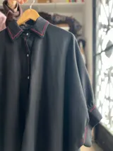 Camisa oversize de lino color negro, con cuello clásico, cierre frontal con botones y detalles de costuras bordadas en color rojo. Presenta mangas anchas y corte holgado.