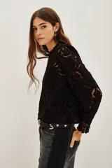 Camisa de encaje negro con cuello alto y mangas largas abullonadas. Presenta un diseño calado con motivos florales y cierre posterior.