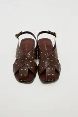 Sandalias planas de cuero sintético marrón, con tiras cruzadas en el empeine y tachas metálicas doradas distribuidas por toda la capellada. Cuenta con tira ajustable en el talón con hebilla.