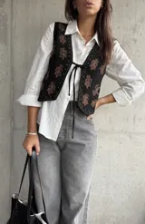 Chaleco negro de gamuza con bordados florales en tonos marrones. Presenta un cierre frontal con lazo y detalles de tachas metálicas en los bordes.