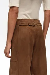 Pantalón tobillero de corte barrel confeccionado en ante de piel caprina, color marrón. Presenta cierre frontal con corchete oculto y cremallera, tiro medio, bolsillos laterales delanteros, pinzas en el delantero y tiras con hebilla en la parte trasera.