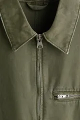 Campera de algodón en color verde militar, con cuello camisero, cierre frontal, un bolsillo superior con cierre y dos bolsillos laterales ribeteados.