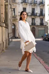 Vestido corto color blanco, de corte holgado con cintura elástica. Presenta escote en V con lazo de ajuste, mangas largas abullonadas con puños elásticos y falda con volados decorados con encaje en el ruedo.