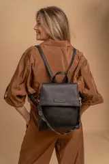 Mochila tipo cartera con tapa, confeccionada en símil cuero color suela. Cuenta con doble compartimento, división interna, cierre de seguridad y bolsillo trasero con cierre. Incluye una correa frontal decorativa con tachas metálicas.