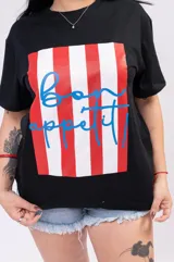 Remera negra de corte oversize con cuello redondo y manga corta. Presenta un estampado frontal rectangular con rayas verticales rojas y blancas, y la inscripción "Bon appétit!" en color azul.
