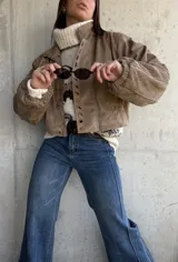 Campera bomber de gamuza color beige, con cierre frontal y detalle de ojales metálicos decorativos en la solapa. Presenta mangas largas con puños elásticos y un corte holgado tipo oversize.