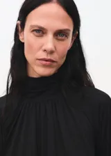 Blusa negra de cuello alto con cierre de botones, diseño recto y mangas largas acampanadas con volantes en los puños. Confeccionada en tejido fluido.
