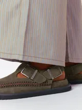 Slipper tipo guillermina confeccionada en gamuza color marrón, con plataforma de goma negra y doble hebilla ajustable. Presenta un pespunte decorativo en el borde inferior y cuenta con interior recubierto en cuero.