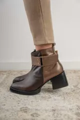 Bota corta de cuero negro con acabado combinado mate y charol. Presenta una tira decorativa con tachas metálicas y hebilla en el tobillo, tacón cuadrado robusto y suela con plataforma.