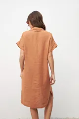 Vestido camisero de manga corta, color tostado/terracota, con cuello solapa y botones frontales. Presenta un bolsillo de parche en el pecho y parece ser de tejido ligero, posiblemente lino o una mezcla.
