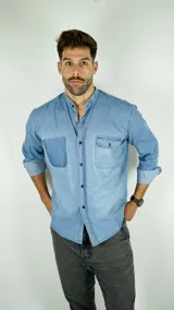 Camisa de jean en tono azul claro con cuello mao y cierre frontal de botones. Presenta un bolsillo frontal con solapa y un detalle de parche en el lado opuesto, con mangas largas arremangadas.