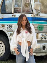 Camisa de lino blanca con mangas largas abullonadas y nudo frontal. Presenta un bordado artesanal en el pecho con diseño de sol y corazón en color rojo.