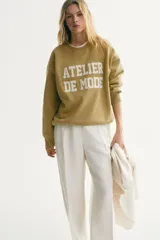 Buzo oversized color beige claro, con cuello redondo, hombros caídos y el texto "ATELIER DE MODE" en relieve con textura de felpa blanca en el frente. Cuenta con puños, cuello y dobladillo en ribete acanalado.