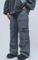 Pantalón cargo técnico gris oscuro, de silueta oversize y tejido liviano. Presenta múltiples bolsillos cargo con solapa en las piernas, uno de los cuales tiene una pequeña etiqueta negra con texto.