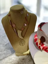 Collar de cadena dorada con múltiples dijes colgantes en tonos rosados, incluyendo piezas de nácar y un dije central en forma de colmillo color crudo, acompañado por un dije circular con diseño de árbol de la vida.