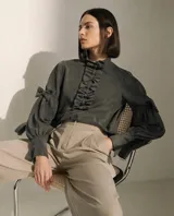 Blusa de color verde oscuro con cuello alto con volados, detalle de lazos frontales y mangas largas abullonadas con lazos decorativos en los codos y puños ajustados.