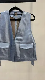 Chaleco corto de efecto metalizado en tono celeste claro, con cuello en V, cierre frontal con un botón a presión y dos bolsillos delanteros con solapa.