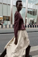 Chaqueta color beige con efecto ante, cuello redondo, manga larga y cierre frontal con cremallera. Presenta un bajo en evasé y un cinturón fino de cuero marrón oscuro con hebilla metálica.