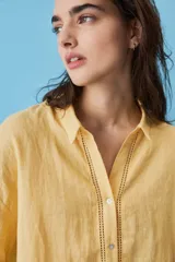 Camisa blanca de lino, con mangas anchas a tres cuartos, cuello clásico, cierre frontal con botones y detalles de calados verticales en la parte central y en los puños.