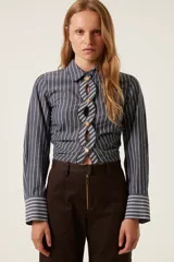 Camisa corta a rayas verticales grises y blancas, con cuello clásico y mangas largas. Presenta un diseño trenzado en el frente con botones dorados y aberturas.