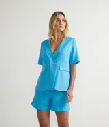 Blazer celeste de lino con escote en V, manga corta, cierre frontal con dos botones dorados y bolsillos de solapa.