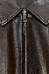 Chaqueta estilo bomber de efecto piel en color marrón oscuro, con cuello solapa, cierre frontal con cremallera, bolsillos delanteros de vivo y bajo acabado en elástico.