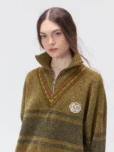 Pullover de punto con textura aterciopelada, diseño oversize, cuello polo con cierre frontal parcial y detalles de pespuntes en contraste. Presenta rayas horizontales en tonos tierra y un parche bordado con la letra M en el pecho.