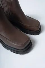 Bota corta tipo Chelsea de cuero negro, con puntera cuadrada y suela track gruesa y dentada. Presenta paneles elásticos acanalados a los lados para facilitar el calce.