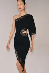 Vestido negro de escote asimétrico con un solo hombro y manga larga ancha. Presenta detalles de encaje negro en el escote, el ruedo asimétrico y un corte 'cut out' en el lateral del torso.