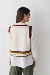 Chaleco de lana color marrón con cuello estilo bebé, cierre frontal con botones grandes y detalle de puntilla en cuello, sisas y bolsillo frontal. Presenta una franja decorativa en color rojo en el ruedo.