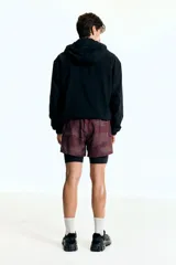 Short deportivo de doble capa, con calza interior. El short exterior es de color borgoña con un estampado sutil de puntos en forma de camuflaje. Posee cintura elástica con cordón oculto y aberturas laterales. La calza interior es negra.