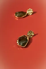 Aros colgantes dorados con forma irregular, engastados con una piedra o resina de color marrón oscuro y verde azulado veteado. El poste superior tiene un diseño orgánico y texturizado.