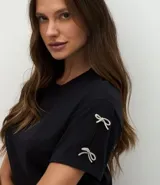 Camiseta blanca de algodón con cuello redondo y mangas cortas, decorada con pequeños lazos brillantes en los laterales de las mangas.