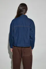 Campera de jean azul oscuro, de corte oversize, con cuello clásico, cierre frontal con cremallera y botones a presión, bolsillos frontales con solapa y mangas largas.
