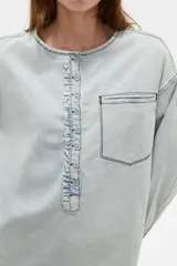 Blusa de jean ligera en color celeste claro, con cuello redondo y cierre frontal parcial con botones. Presenta un diseño con volados a lo largo de la botonera, bolsillo ojal en el pecho, y mangas largas abullonadas con puños ajustados y fruncidos.