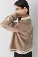 Cardigan de punto color crema con cuello redondo y cierre frontal de botones. Presenta detalles bordados de motivos celestiales y marinos, con terminaciones en tejido acanalado a rayas azules en cuello, puños y ruedo.