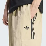 Pantalón cargo Adidas de corte holgado, color beige, confeccionado en tejido antidesgarre. Presenta las icónicas tres rayas laterales en negro, bolsillos cargo en los laterales y logo del trifolio bordado en la pierna izquierda.